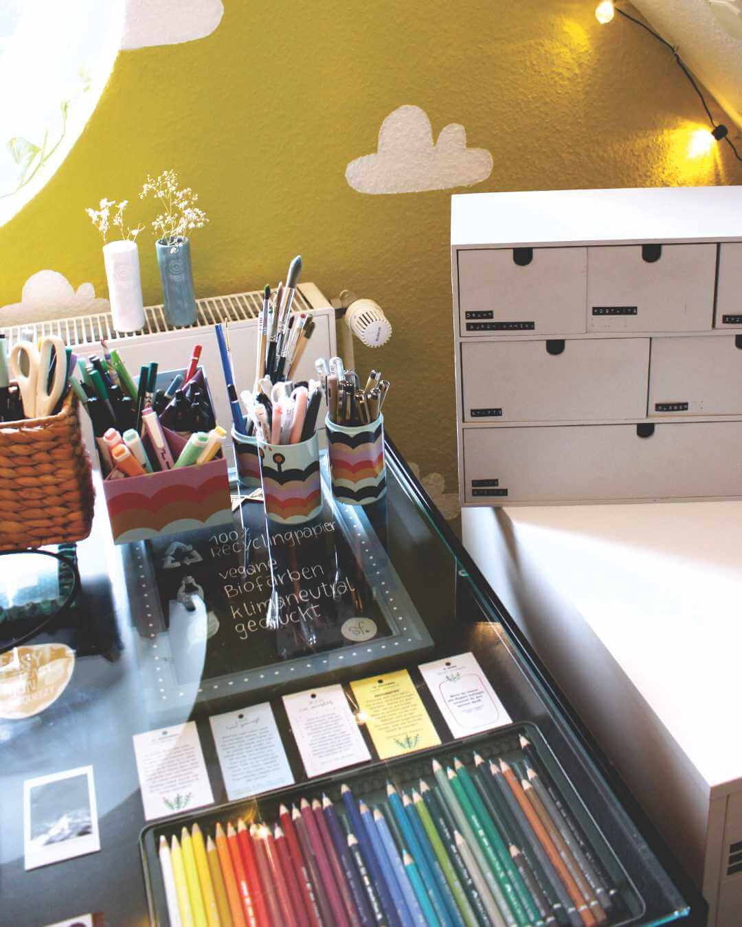 schreibtisch-mit-stiften-und-boxen-vor-gelber-wand-mit-weissen-wolken-Atelier-Sintje-Tramm